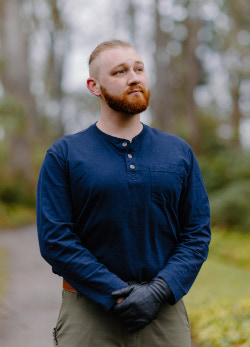 Stoic headshot of man with red beard and pony tail in the woods.
